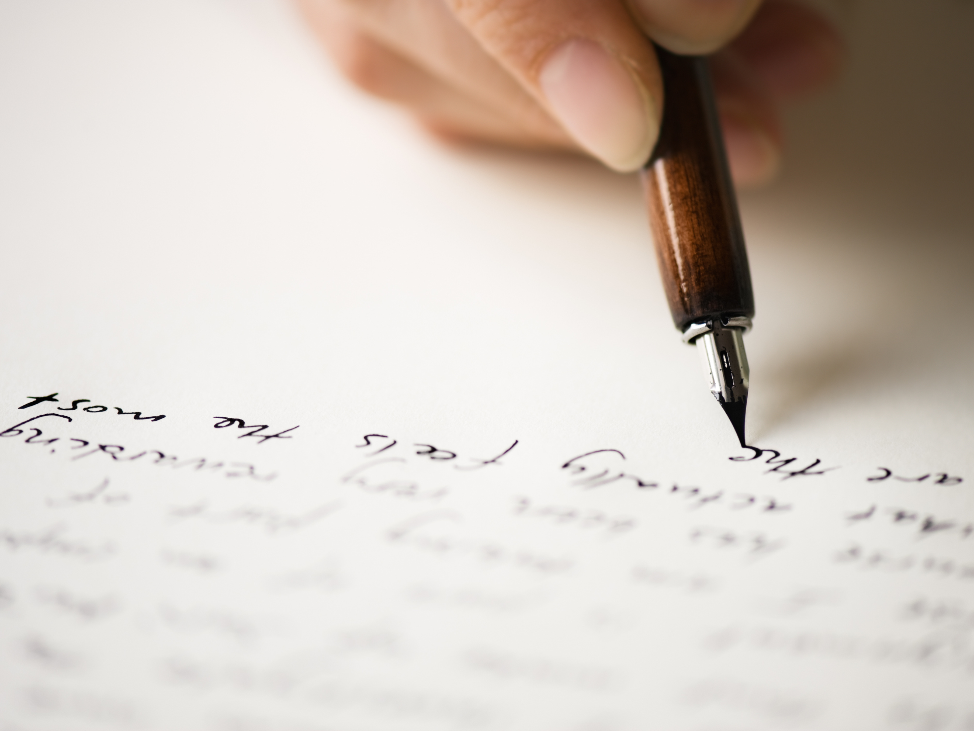 A close-up image of a hand holding a wooden fountain pen, writing in elegant, cursive script on white paper. The focus is on the pen tip and the text being written, with the background softly blurred. The hand appears to be steady, and the ink is a dark, rich black.